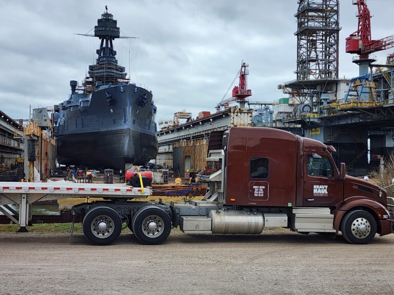 Heavy Haul Solutions flatbed trailer and semi tractor parked at industrial shipyard with naval vessel under construction in background