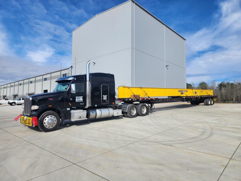 Semi tractor hauling yellow beam parked at facility yard with warehouse building in background.