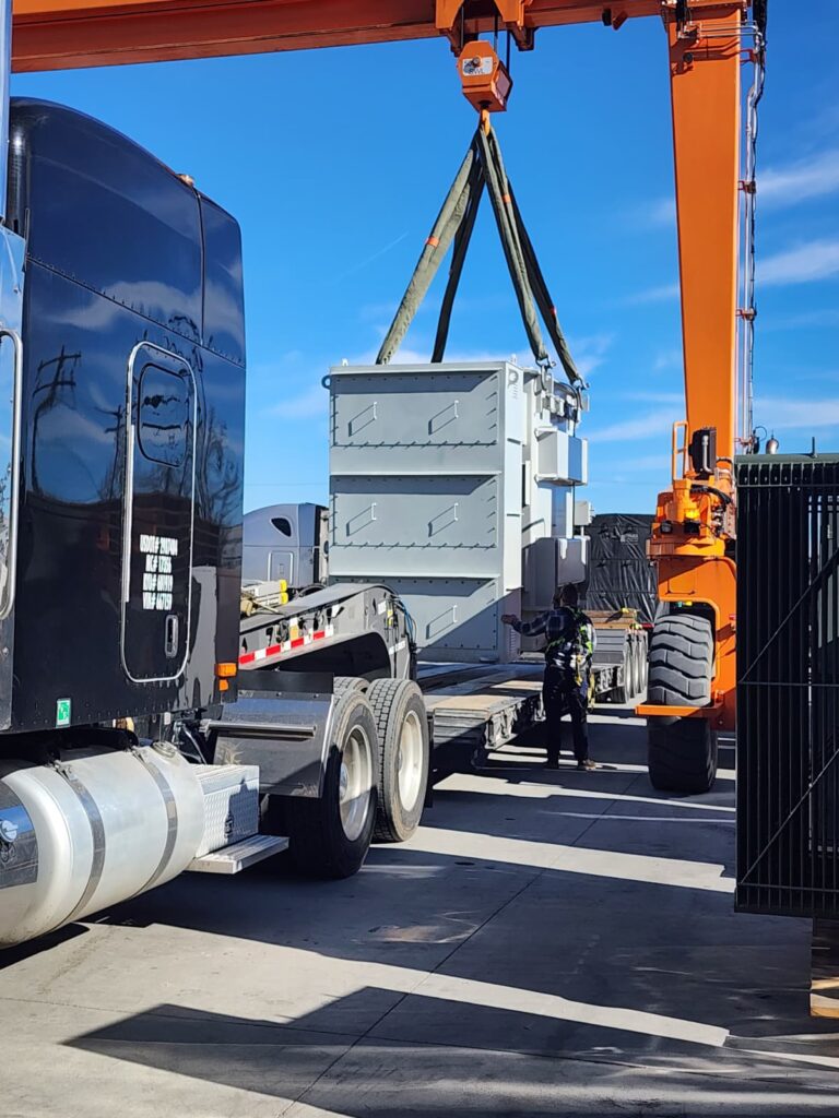 Orange mobile crane with spreader bar hoisting white industrial transformer trailer above parked semi truck at industrial yard.