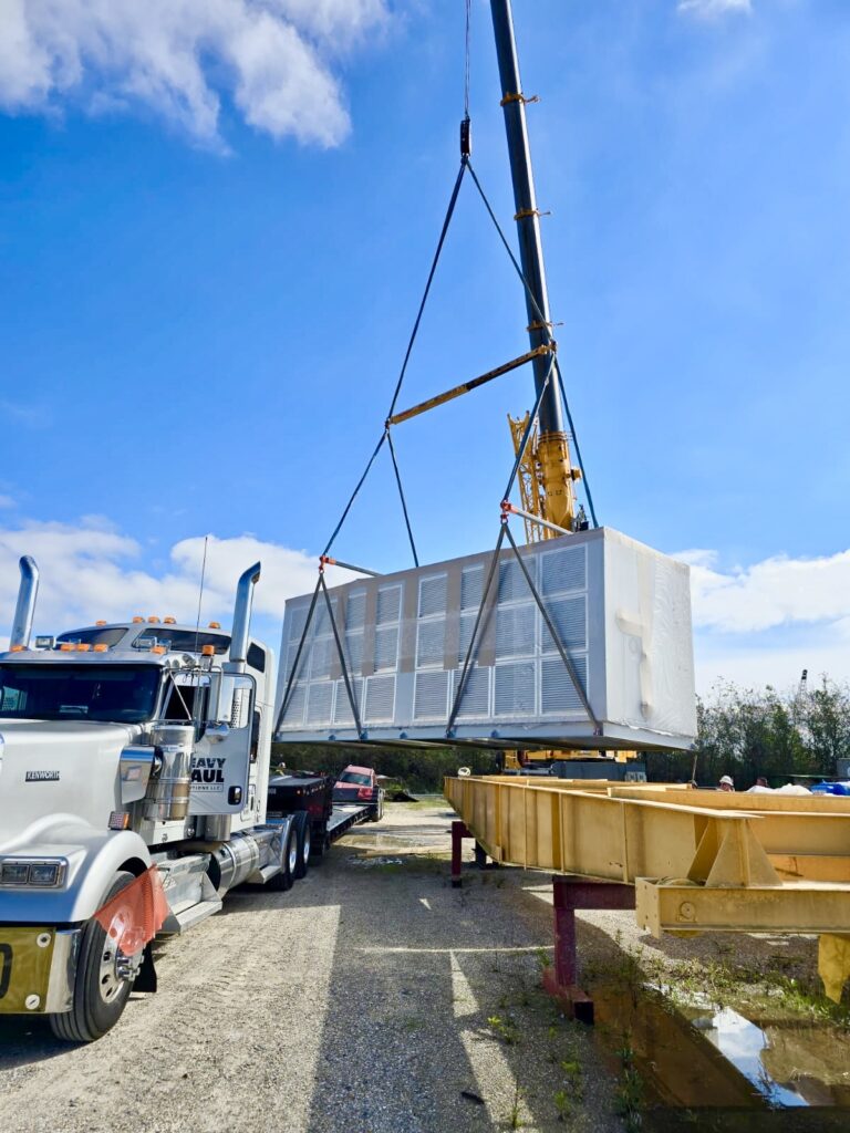 Semi truck parked at yard with crane lifting large white industrial unit onto RGN trailer under clear sky