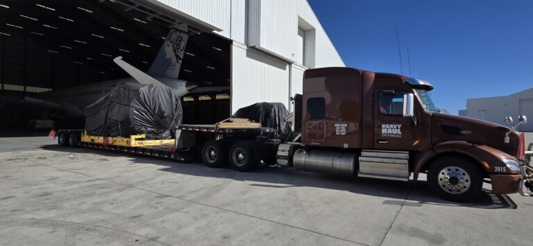 Heavy Haul Solutions tractor and step deck trailer parked outside a hangar with oversized aircraft fuselage wrapped in protective covering on the trailer.