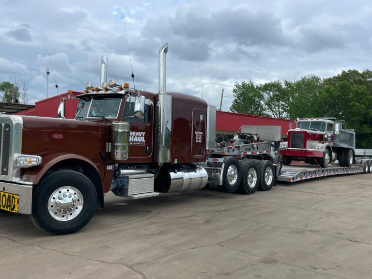 Semi tractor with stepped trailer parked at yard, carrying red dump truck on flatbed; another truck visible in background.
