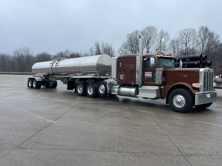 Heavy Haul Solutions truck hauling a tanker trailer parked on concrete lot with bare trees in background.