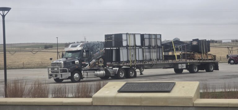 Semi tractor with flatbed trailer carrying large industrial units parked at a terminal, rural landscape in background.