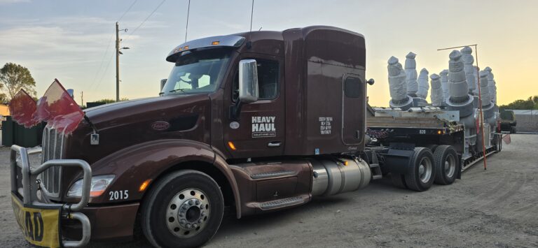 Heavy Haul Solutions semi tractor with maroon cab and flatbed trailer carrying large wrapped industrial units in a yard at dusk.
