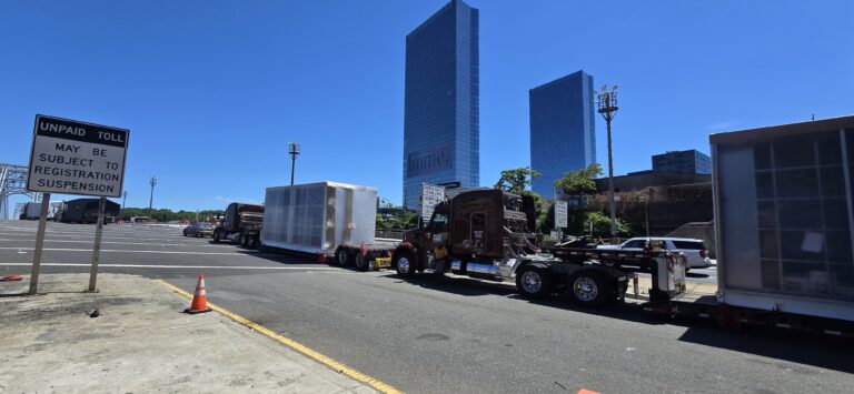 Heavy haul trucks parked in a paved yard with tall buildings in background; one truck carries wrapped cargo on a flatbed trailer.