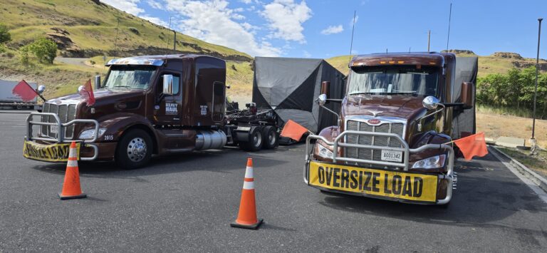 Two semi trucks with oversized load signs parked on a roadway with traffic cones and safety equipment visible, hillside in background.