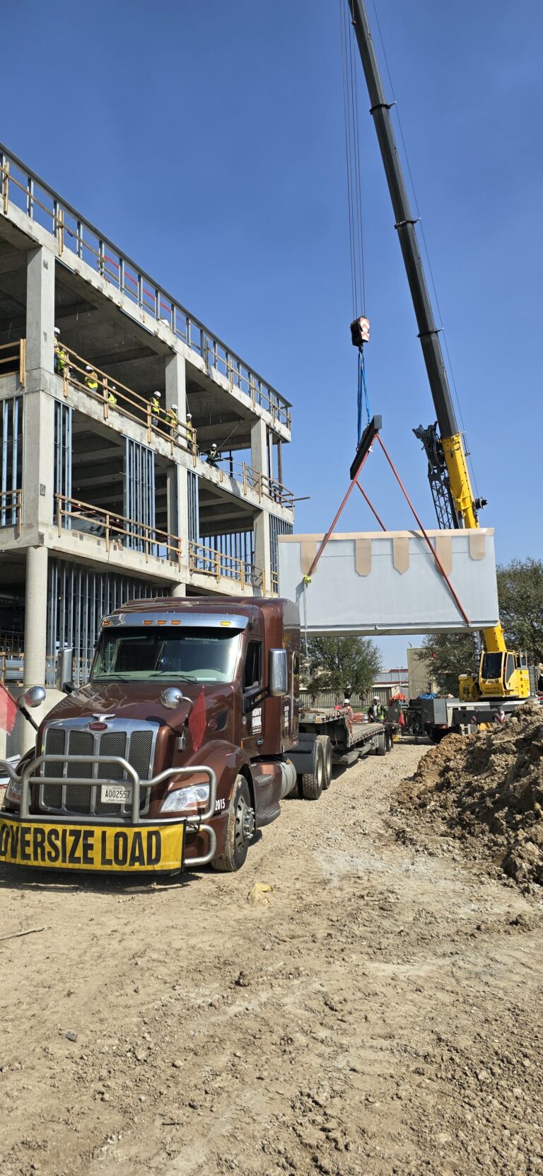 Semi truck with oversized load sign parked at construction site with crane unloading modular building component