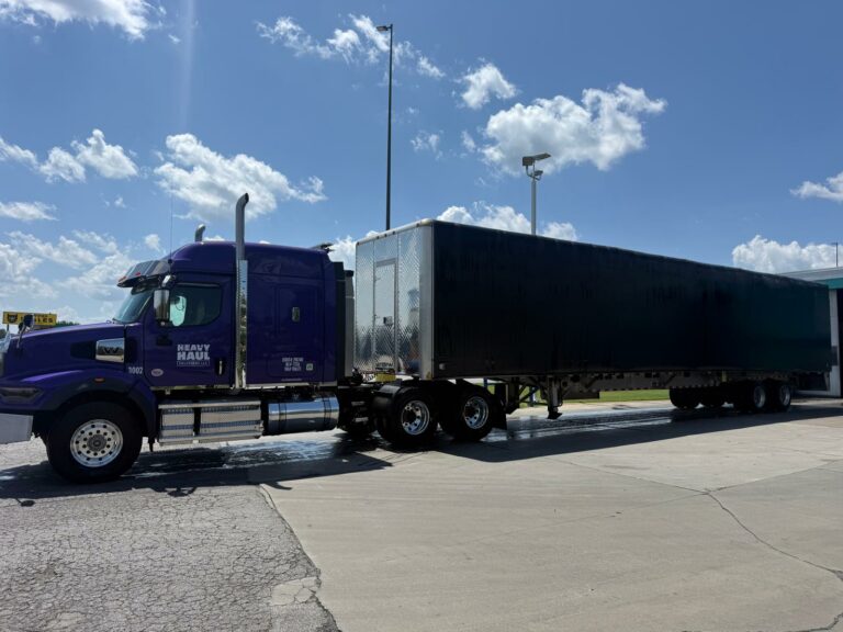Heavy Haul Solutions semi tractor with Conestoga trailer parked at a terminal under blue sky with white clouds.
