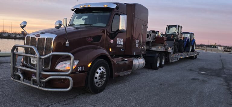 Heavy Haul Solutions semi tractor with landoll trailer carrying tractor equipment, parked at waterfront facility at dusk.