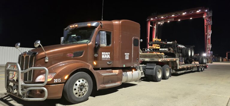 Semi tractor with Heavy Haul Solutions branding parked next to enclosed car hauler trailer at night in yard.