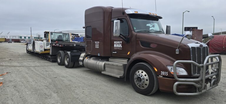 Semi tractor with Heavy Haul Solutions branding parked at yard, pulling equipment on RGN trailer.