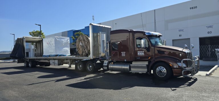 Heavy Haul Solutions semi tractor with flatbed trailer carrying large cylindrical industrial equipment and machinery parked at terminal.