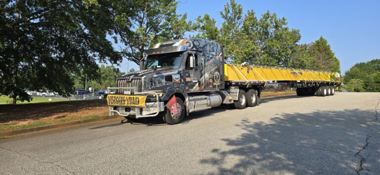 Semi tractor with flatbed trailer parked at a facility with trees, a second yellow flatbed trailer visible in background.