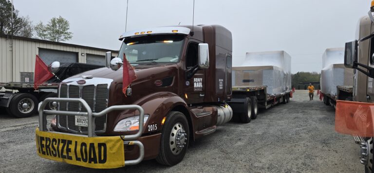 Heavy Haul Solutions semi tractor with oversize load parked in yard with other trailers and workers in background.