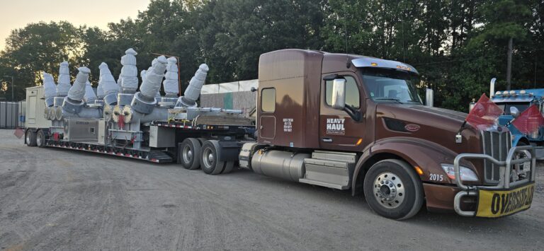 Semi tractor with flatbed trailer carrying large industrial units wrapped in white coverings, parked in yard with trees and equipment in background.
