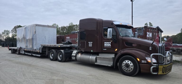 Semi tractor with Heavy Haul Solutions branding parked at a yard with a flatbed trailer carrying a large white industrial unit.