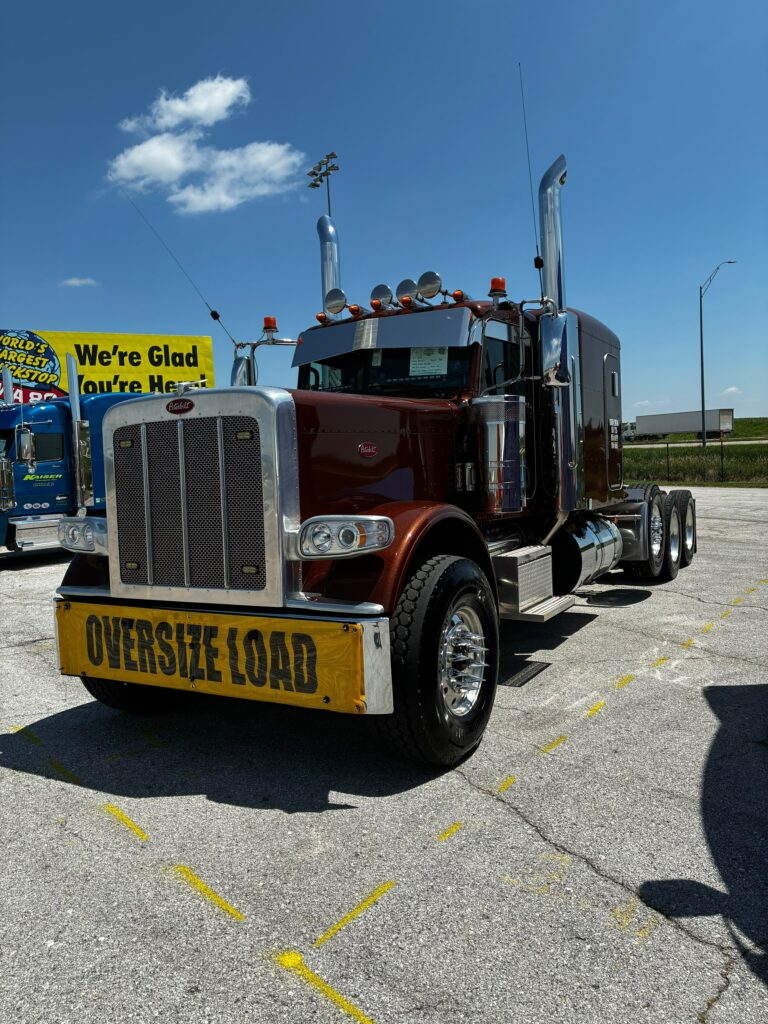 Semi tractor with red cab and chrome details parked at an event, displaying yellow oversize load sign on front bumper. Walcott Truckers Jamboree Super Truck Beauty Contest