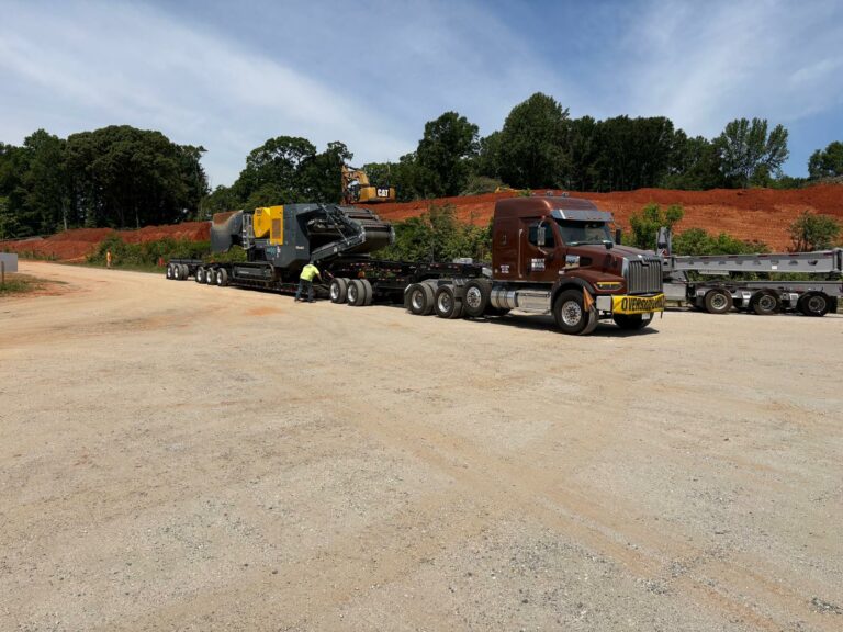 Heavy haul truck with black lowboy trailer parked at dirt yard with red dirt pile and trees in background.