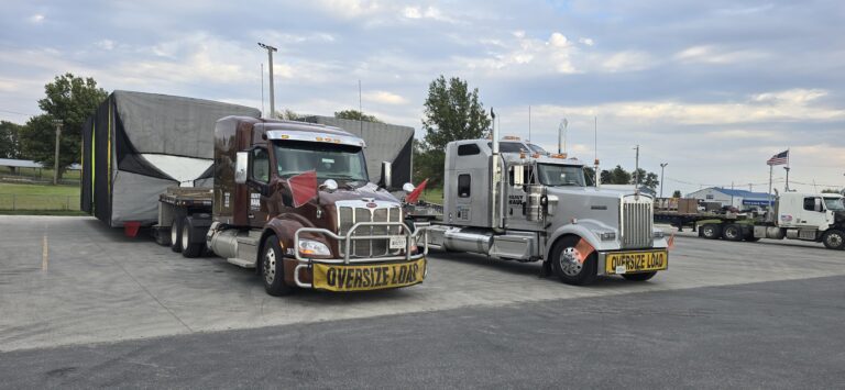 Two tractor-trailers with oversize load banners parked in a lot, one hauling an enclosed cargo box, the other a white cab with equipment visible.