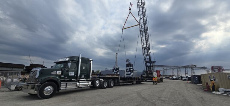 Semi tractor with step deck trailer parked at industrial facility with crane loading it.