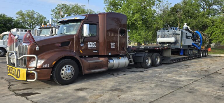 Heavy Haul Solutions semi tractor with brown cab parked next to step deck trailer carrying industrial equipment at yard with other trucks in background
