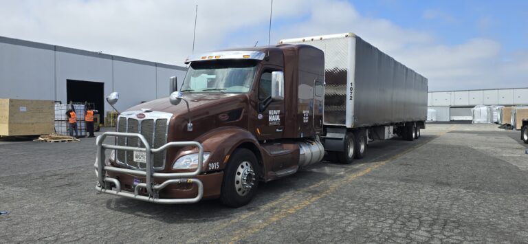 Semi tractor with enclosed trailer parked at industrial facility with workers and buildings visible in background.