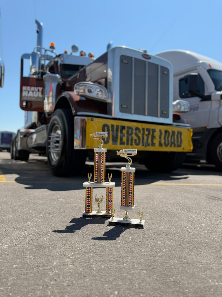 Trophy displayed on pavement in front of Heavy Haul Solutions truck with oversize load banner parked at terminal. Walcott Truckers Jamboree Super Truck Beauty Contest