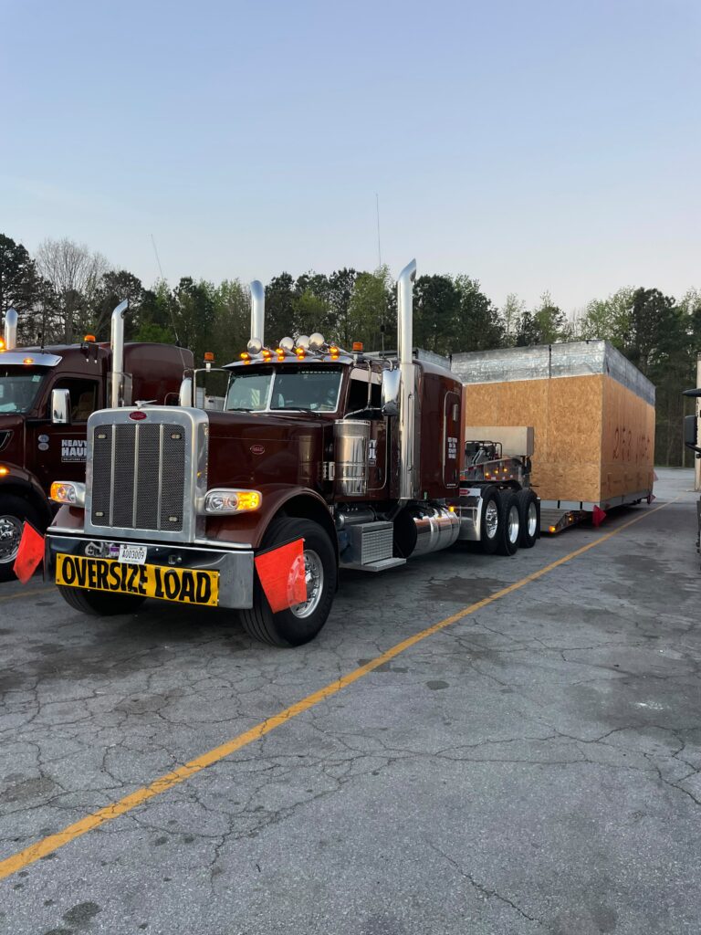Semi tractor with tall chrome stacks pulling enclosed step deck trailer marked with oversized load signs, parked at terminal.