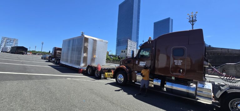 Semi tractor with brown cab parked next to white enclosed trailer in industrial yard with city skyline in background.