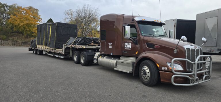Semi tractor with flatbed trailer loaded with industrial equipment and cargo tarped and secured at an outdoor yard, other trailers visible in background.