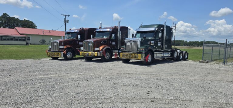 Three heavy-haul semi tractors with oversize load banners parked at a yard with farm buildings and fields in background.