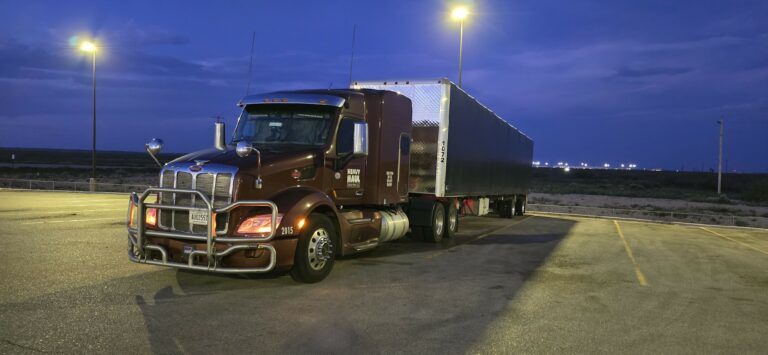 Semi tractor with enclosed trailer parked in lot at dusk under overhead lighting.