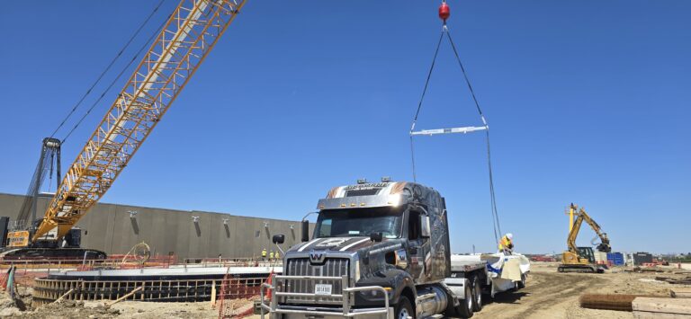 Semi tractor and flatbed trailer parked at industrial site with large mobile crane, excavator, and construction materials visible.