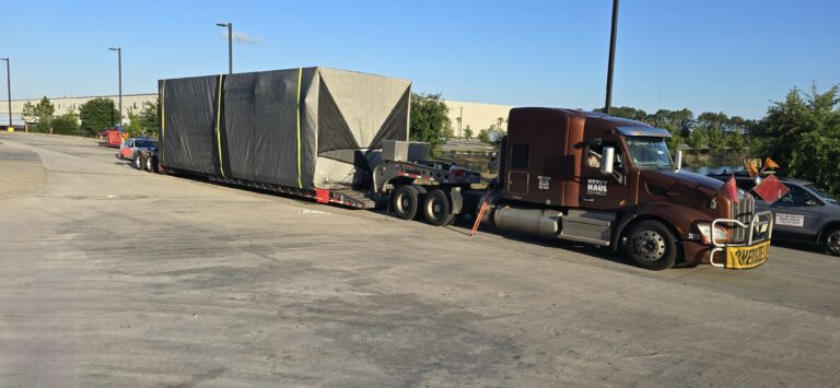 Semi tractor pulling RGN lowboy trailer with large wrapped industrial equipment parked in lot with other trucks nearby.