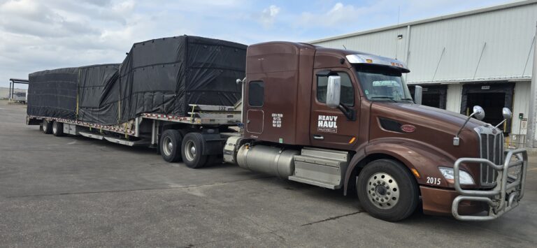 Semi tractor with brown cab parked beside flatbed trailer with black tarp covering cargo at industrial facility.