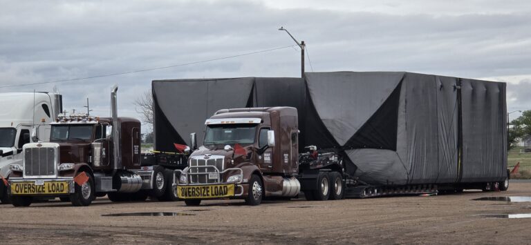 Two semi tractors with oversize load placards parked with a large covered lowboy trailer carrying oversized industrial cargo.