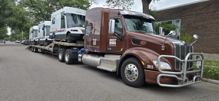 Semi tractor with flatbed trailer carrying Oshkosh NGDVs mail trucks, parked on residential street near brick building.
