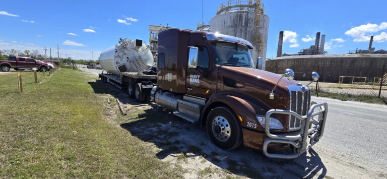 Heavy Haul Solutions truck hauling a step deck trailer with a tank, parked at industrial facility with storage tanks in background.
