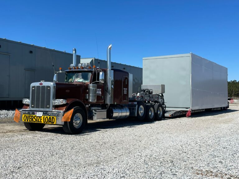 Semi tractor with oversize load marker hauling a large white enclosed trailer on a gravel lot near industrial buildings under clear sky.