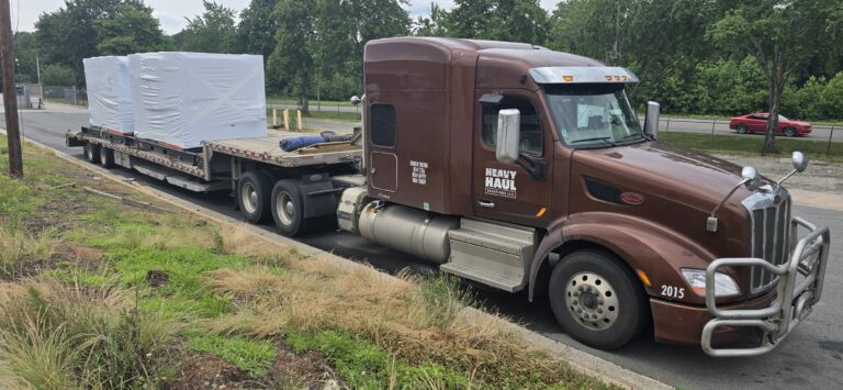 Heavy Haul Solutions semi truck with brown cab parked on roadside pulling flatbed trailer loaded with large white-covered cargo and equipment.