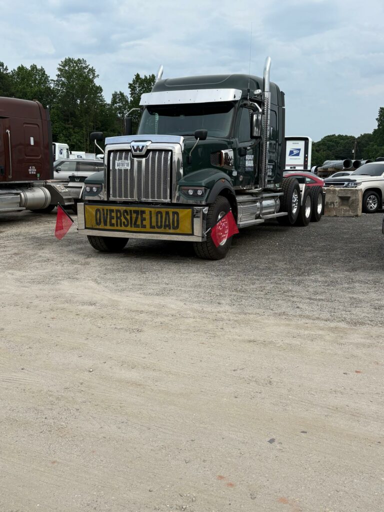 Semi tractor with oversize load sign parked at facility, other vehicles visible in background