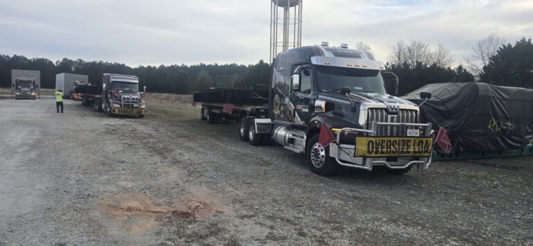 Heavy haul trucks parked in a yard, including a lowboy trailer with oversized load marked with yellow warning signs, surrounded by trees and a water tower.