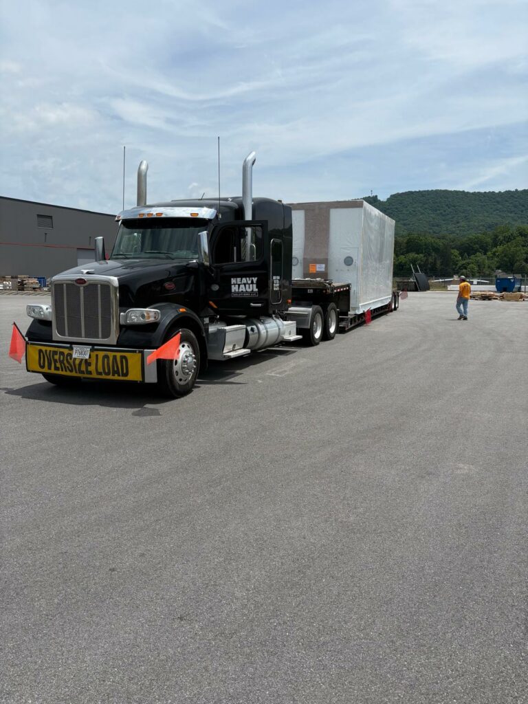 Semi tractor with oversize load sign parked at yard with RGN trailer, worker visible nearby, forested hills in background.