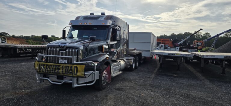 Semi tractor with flatbed trailer parked at heavy haul yard with other equipment and vehicles visible in background.