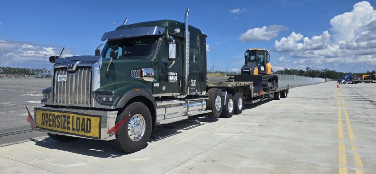 Heavy Haul Solutions semi tractor with step deck trailer carrying construction equipment parked in industrial yard, oversize load sign visible.