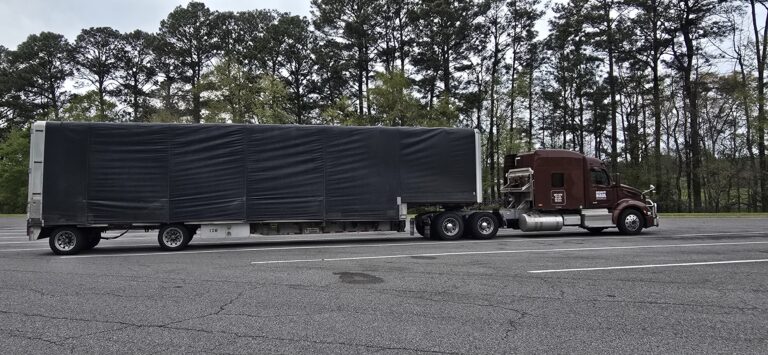 Semi tractor pulling enclosed black trailer on highway with pine trees in background.