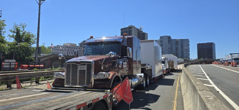 Semi tractors hauling oversized modular units trailer on urban roadway with city buildings in background.