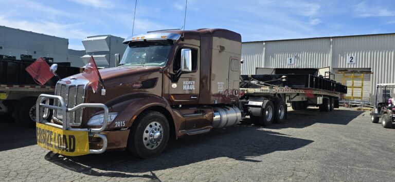 Semi tractor with Heavy Haul Solutions branding parked at yard with flatbed trailer and industrial equipment visible in background.
