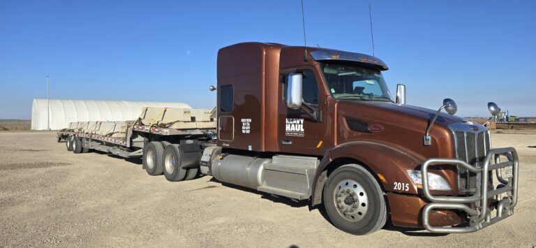 Semi tractor with brown cab parked beside flatbed trailer carrying white cylindrical tank in desert yard.